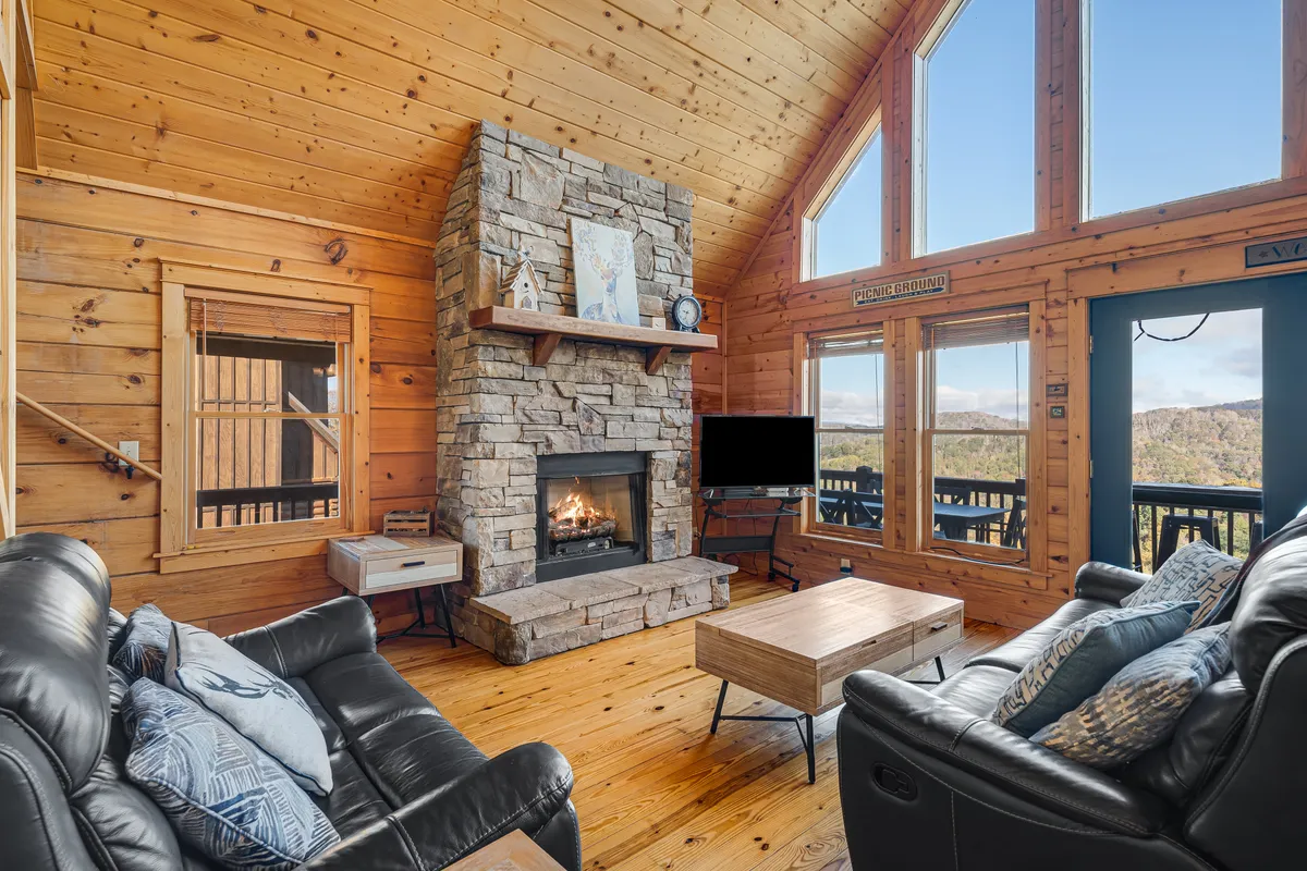 A welcoming living room with a rustic stone fireplace, exposed wooden beams, ample cushioned seating, and expansive windows that let in natural light and showcase the mountains beyond. The space exudes comfort, perfect for movie nights or sharing stories by the fire.