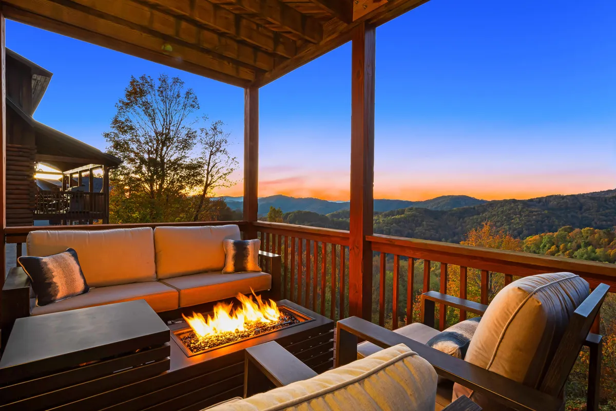 A cozy fire pit area with comfortable seating against a backdrop of rolling mountains and a glowing sunset sky. The arrangement invites guests to gather for warmth, conversation, and s’mores under the stars.