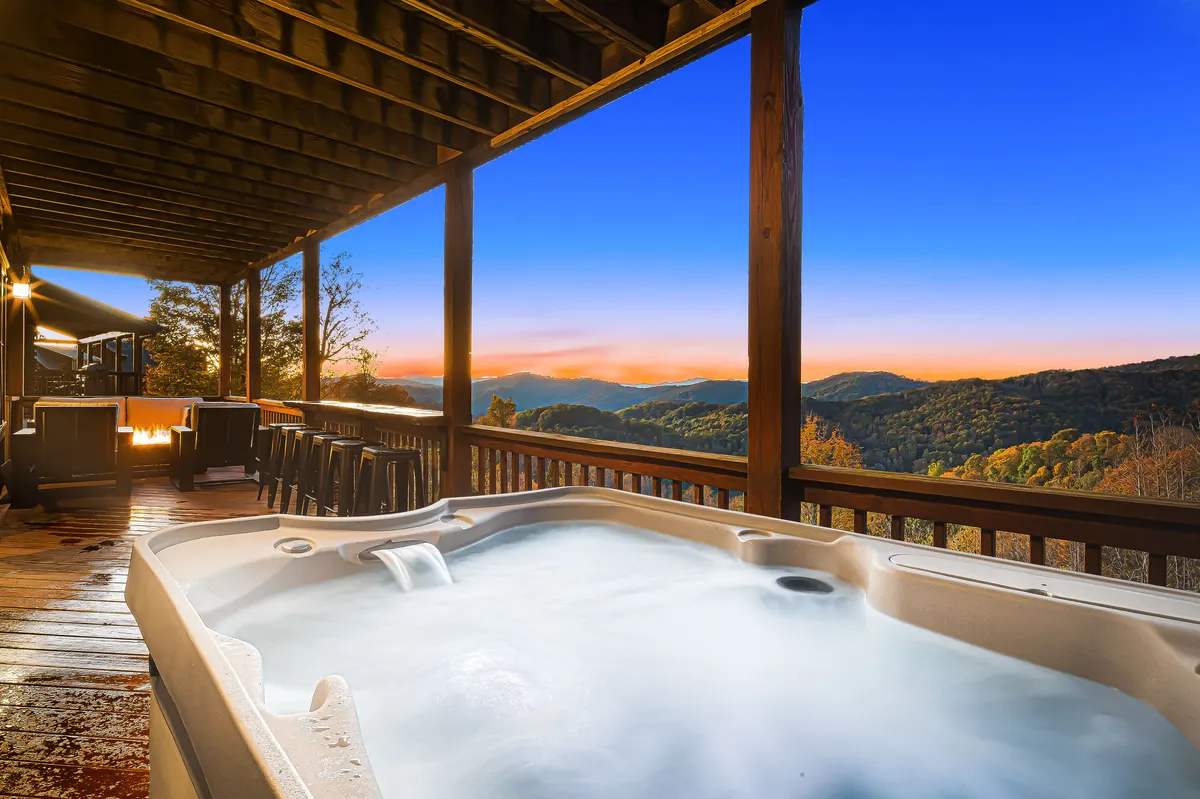 The image shows an inviting outdoor deck overlooking the rolling Blue Ridge Mountains, with a bubbling hot tub as the focal point. Comfortable seating surrounds the area, perfect for gatherings or quiet evenings, while the expansive mountain view stretches into the distance, bathed in the warm glow of sunset.