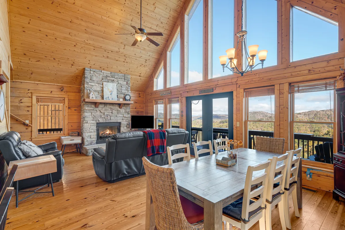 A spacious living area featuring a grand stone fireplace as its centerpiece, flanked by large windows that frame breathtaking mountain views. A comfortable seating area with plush sofas and a dining table create an inviting setting for gatherings, relaxation, and enjoying the changing colors of the sunset.