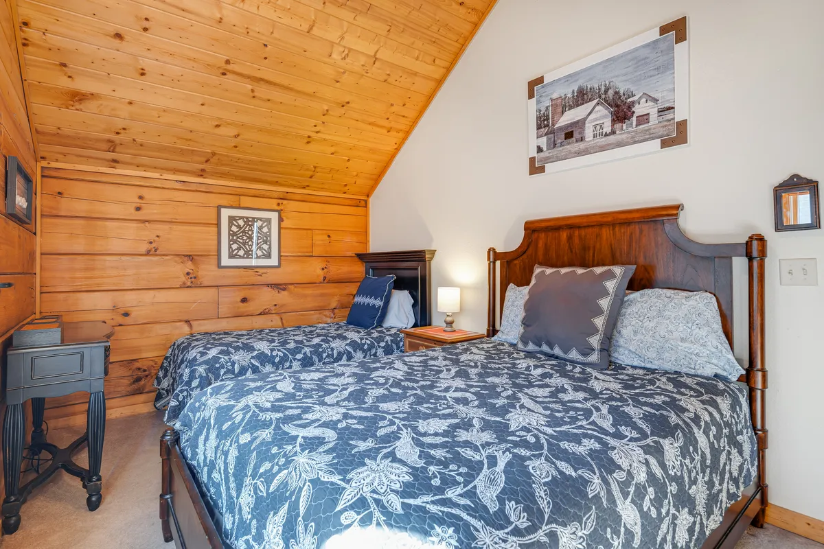 A guest bedroom with two beds adorned with detailed bedspreads, warm wood paneling, framed artwork, and a nightstand with a lamp in between. The room feels intimate and perfect for friends or children sharing the space, inviting laughter and camaraderie.