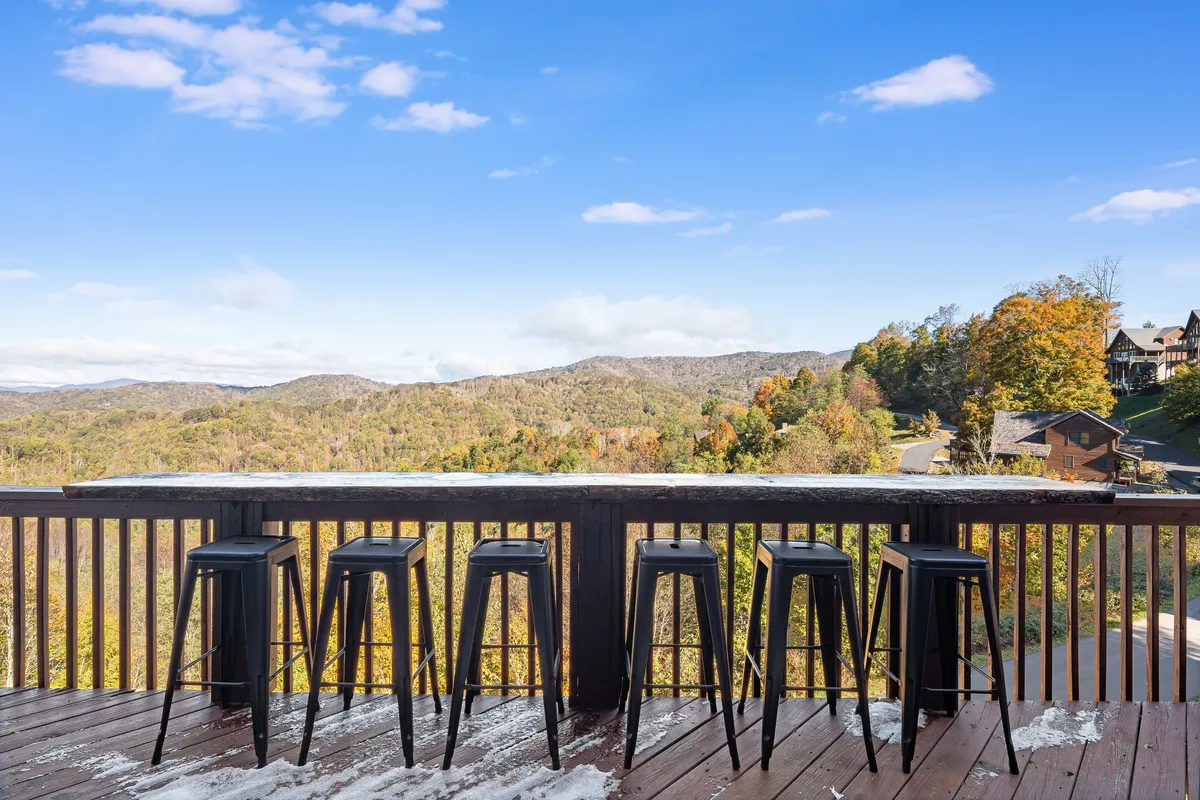 A deck with bar stools and railing frames a majestic mountain view. The setting suggests enjoying a drink with friends while watching the sunset over the peaks.