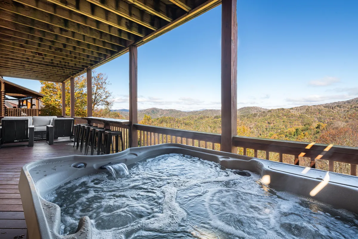 An outdoor hot tub sits on a spacious deck with stunning mountain views. Comfortable seating surrounds the hot tub, making it an idyllic spot for relaxing, socializing, and soaking up the tranquil scenery, day or night.