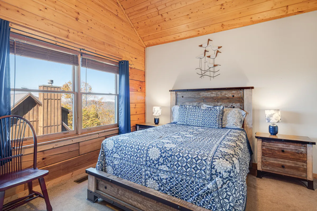A serene bedroom featuring a bed with blue patterned bedding, a large window filling the room with natural light, and wooden accents that evoke a tranquil mountain vibe. The space feels restful and well-appointed for relaxation.