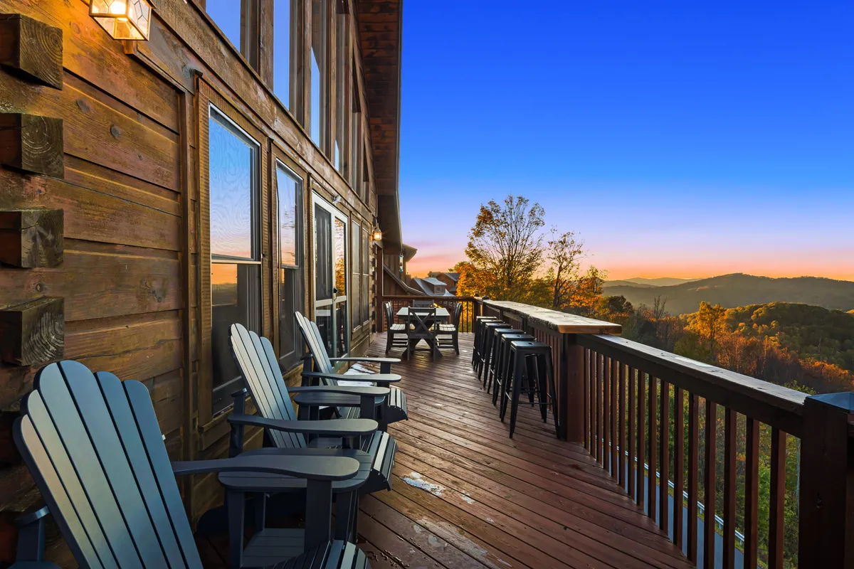 A wooden deck with Adirondack chairs and bar stools faces a sweeping mountain panorama, with a sunset sky painting the scene in warm hues. The space invites socializing, enjoying drinks, and appreciating nature's beauty with friends.