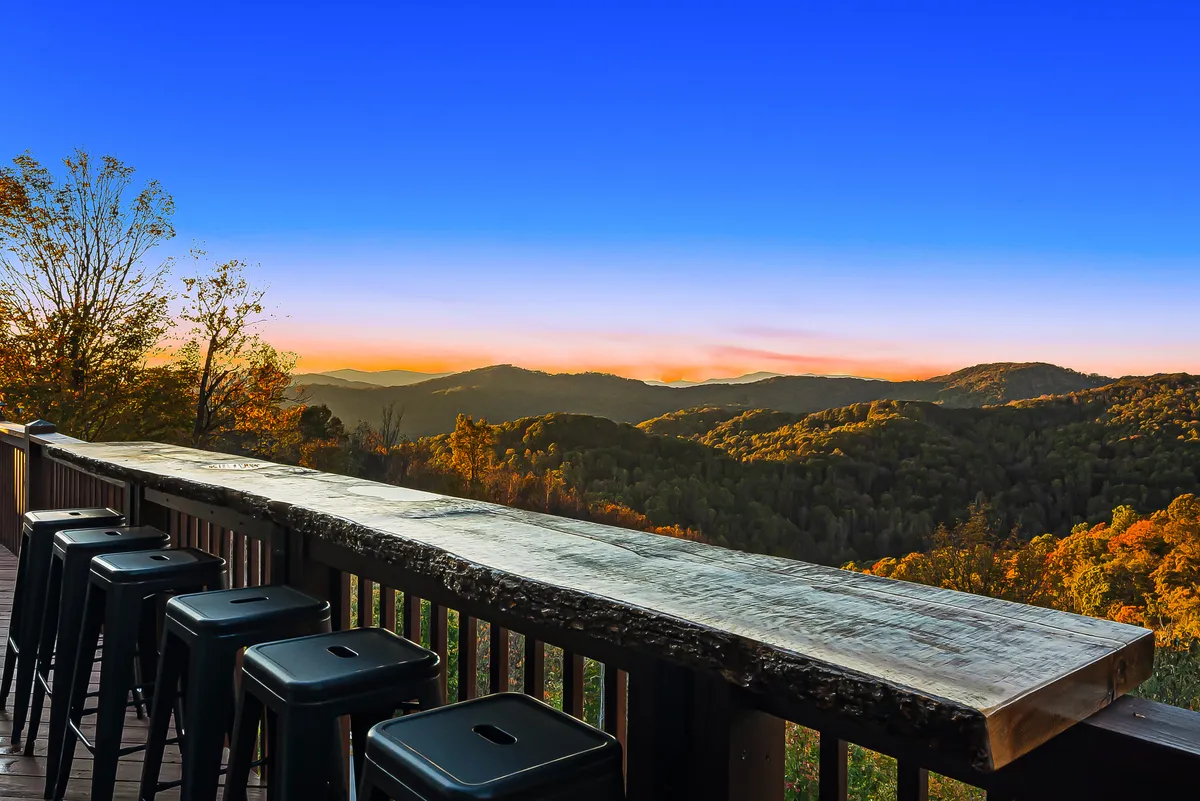 A rustic outdoor bar with metal stools overlooks distant mountains and a vivid sunset sky. The setting is designed for evening cocktails, social gatherings, and soaking in the panoramic view.