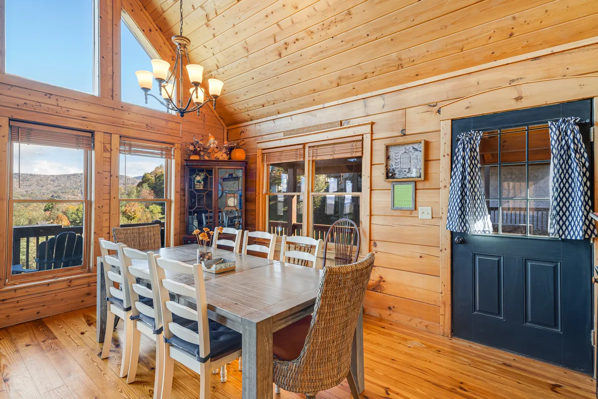 This image features a large wooden dining table positioned beneath a statement chandelier, surrounded by wooden walls and set next to expansive windows. The orientation of the table and windows promises sunset views and a welcoming space for shared meals or game nights.