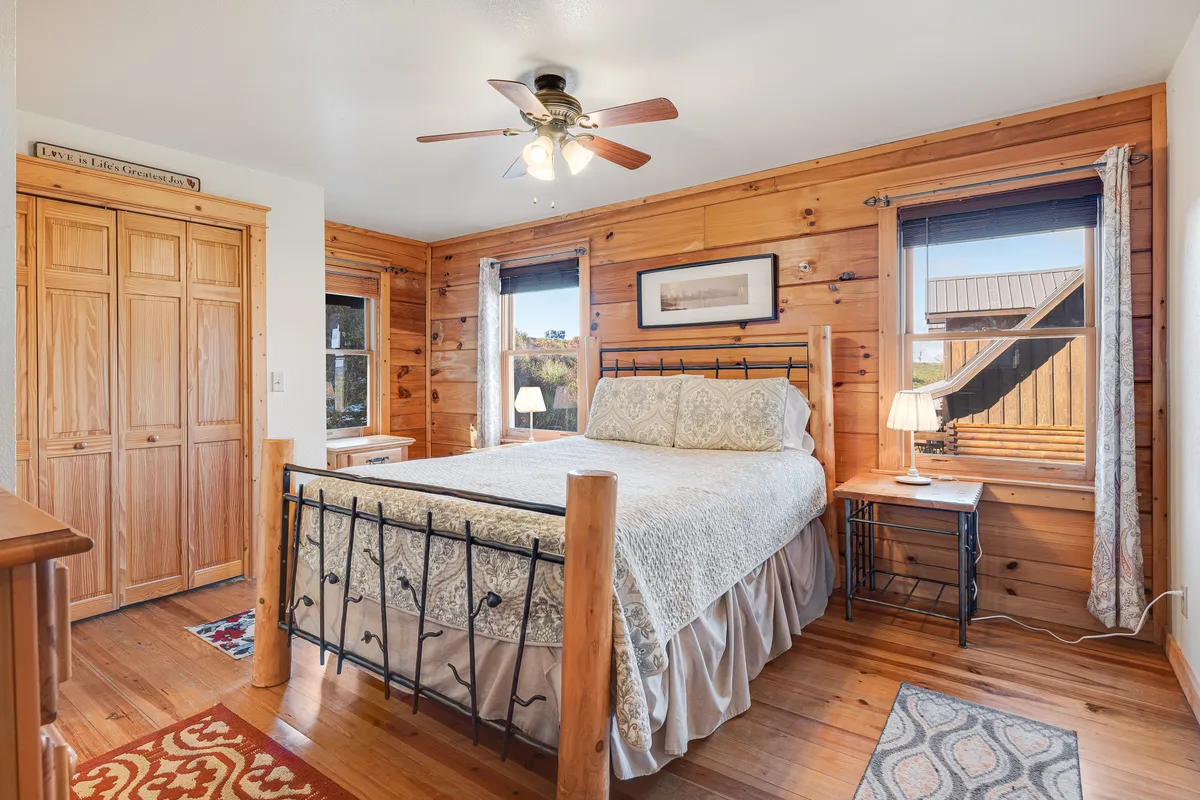 A tranquil bedroom featuring a cozy bed with a neutral bedspread, wooden walls, large windows letting in natural light, a nightstand, and a lamp. The scene exudes warmth and comfort, inviting relaxation and rest after a day in the mountains.