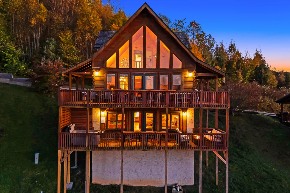 A spacious deck bordered by large glass windows overlooks a surrounding forest, with comfortable outdoor seating arranged for conversation and relaxation. The setting sun casts a golden glow over the wooden deck, hinting at opportunities for gatherings, barbecues, and sunset views.
