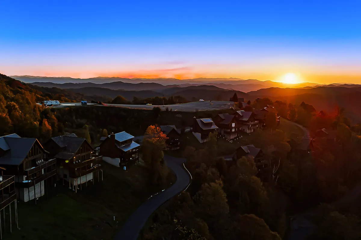 A sweeping view at sunset captures a mountain range with several cozy cabins nestled along a winding road. People can be seen hiking and relaxing on porches, soaking in the natural beauty. The warm tones of the sky and serene landscape evoke a true mountain adventure atmosphere.