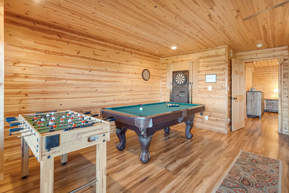 This interior shot spotlights a vibrant game room featuring a foosball table, pool table, and dartboard. The cozy rug and casual seating area create an inviting space for friendly competitions and socializing with friends after a day of outdoor adventure.